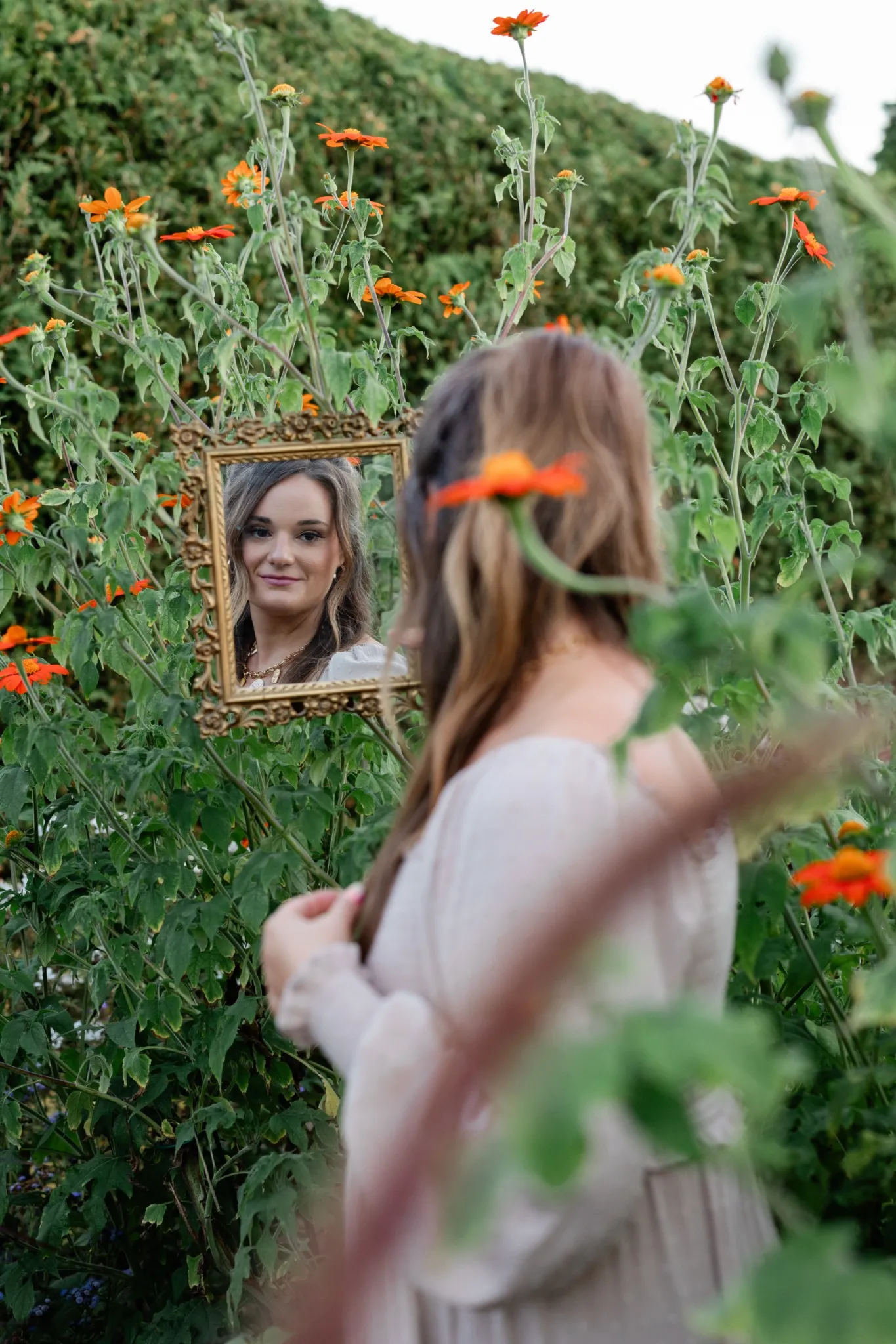 Woman standing among orange flowers, gazing at her reflection in a framed vintage mirror.