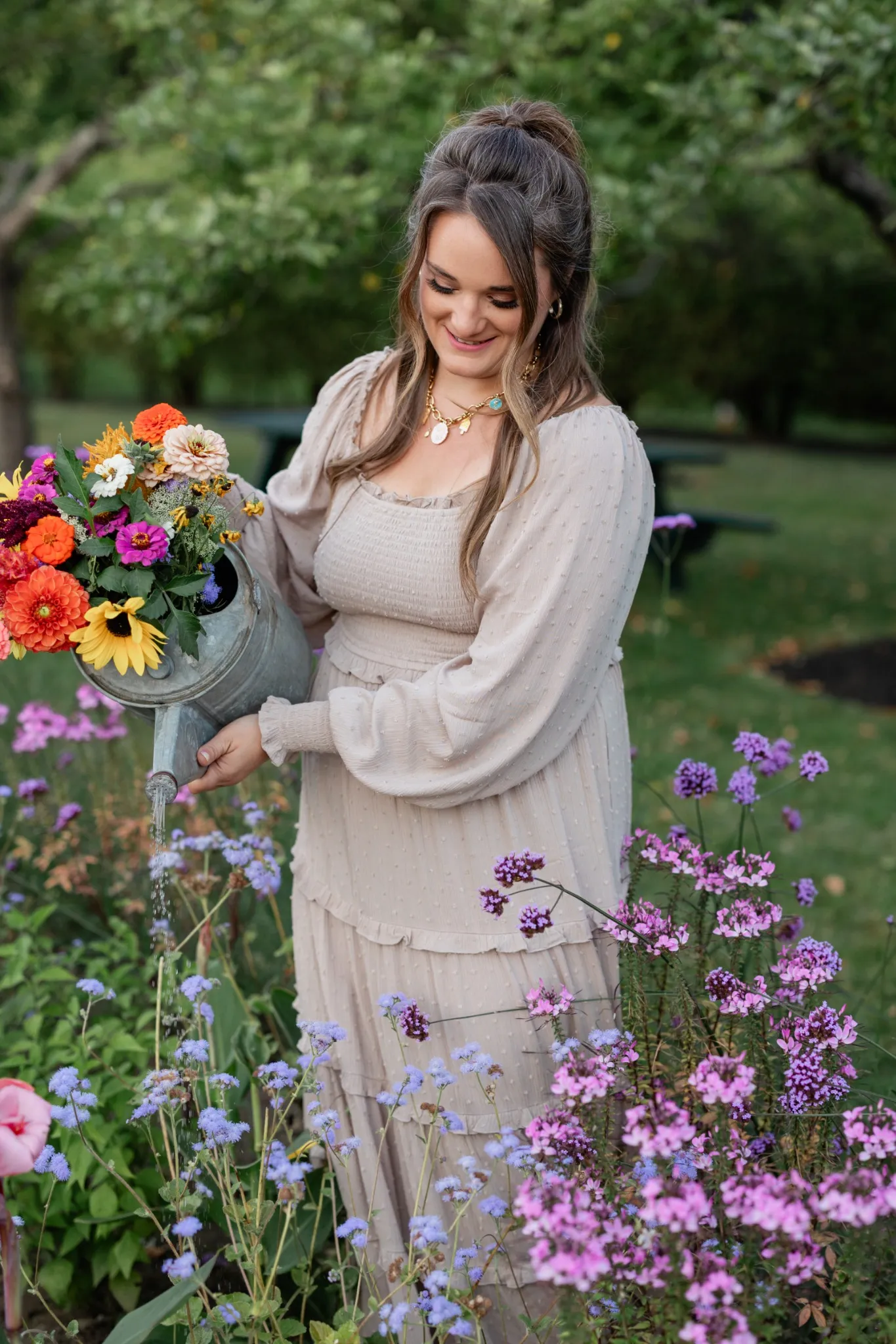 A smiling woman in a light dress waters a variety of colorful flowers in a lush garden.