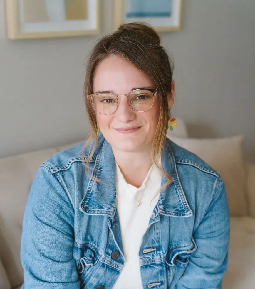 A woman in a denim jacket and glasses smiles warmly while sitting indoors.