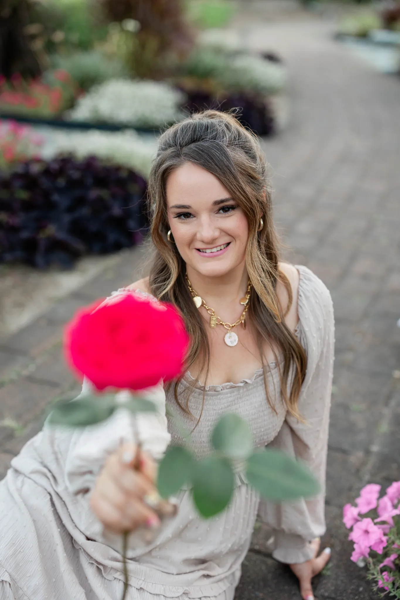 A woman dressed in a light-colored dress offers a bright pink rose to the camera, sitting on a garden path.