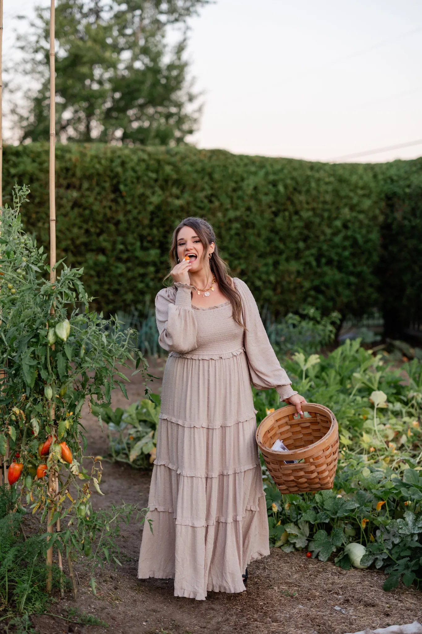 A woman in a flowing dress holds a basket and eats a small tomato while standing in a garden.