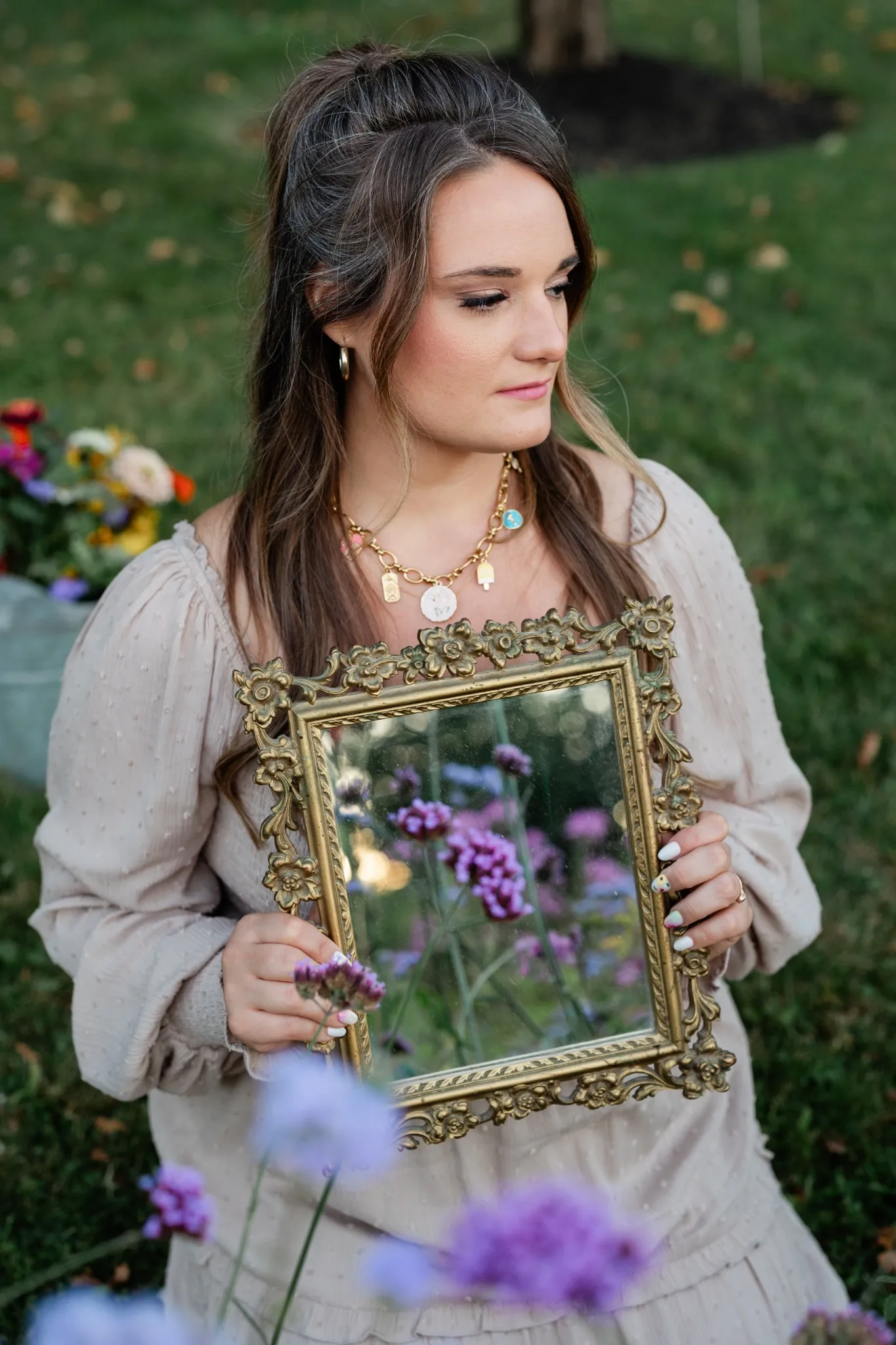 A woman in a garden holds a vintage-style mirror reflecting purple flowers.