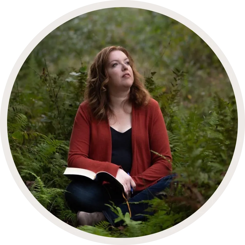 A woman in a red cardigan and black top sits thoughtfully in a forest, holding an open notebook.