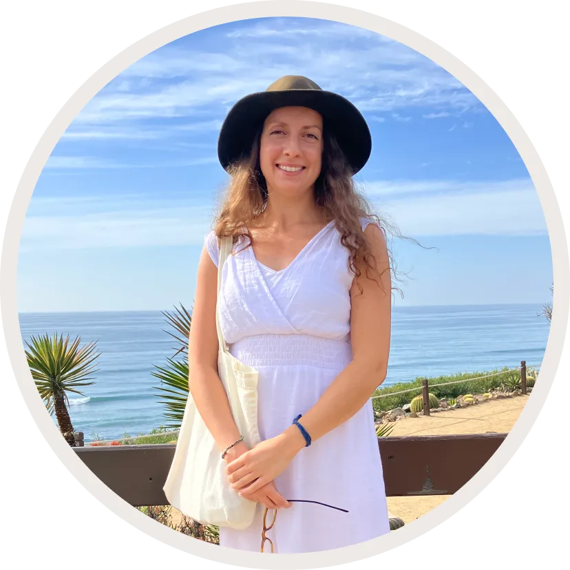 A woman in a white summer dress and wide-brimmed hat standing by a fence with a view of the ocean in the background.