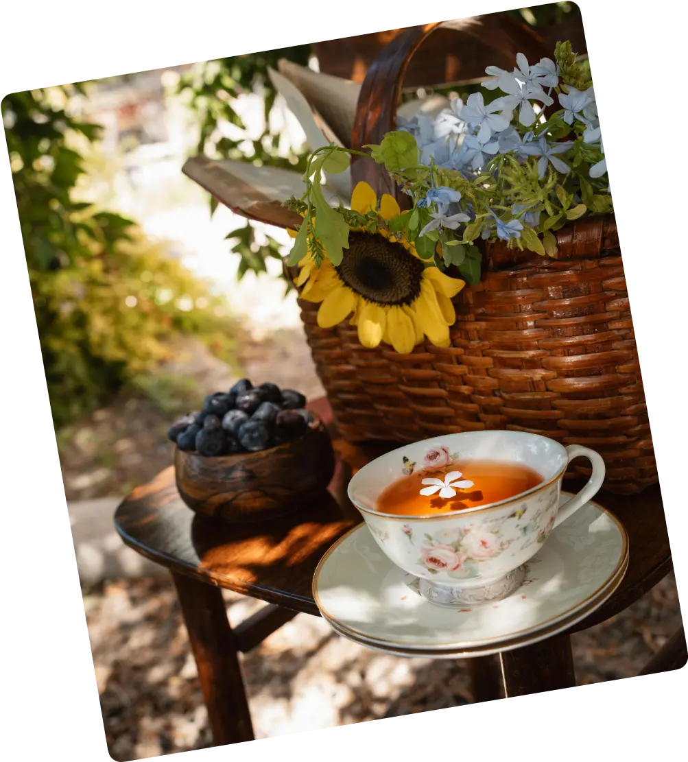 A tea cup on a saucer with a basket of flowers and a bowl of blueberries placed outdoors in a garden setting.